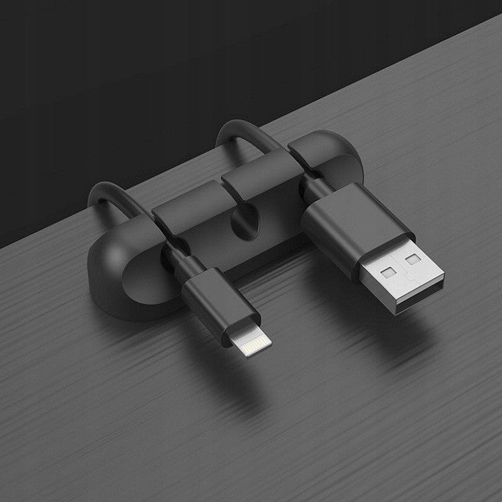 Clemă organizator cabluri din silicon negru lipită pe birou de lemn, fixând cablu USB și cablu audio