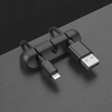 Clemă organizator cabluri din silicon negru lipită pe birou de lemn, fixând cablu USB și cablu audio